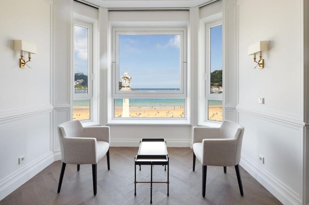 Best Boutique Hotels in San Sebastian 16 Two white armchairs and a small table are arranged in a bright, elegant room with large windows overlooking a sandy beach—capturing the coastal charm found in boutique hotels in San Sebastian, with a decorative clock and people visible outside.