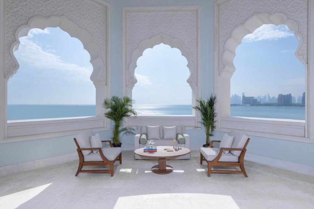 Where to Stay in Doha, Qatar: Transit, City & Beach Hotels 6 Elegant balcony with arched windows overlooking the sea and city skyline, ideal for those seeking where to stay in Doha. Features white cushioned seating, a round coffee table, two potted plants, and a serene atmosphere with blue sky and calm water.