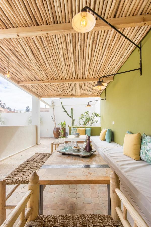 12 Charming Budget Riads in Marrakech 21 A bright, airy patio reminiscent of budget riads in Marrakech, with a bamboo roof, green accent wall, built-in bench seating with colorful cushions, a wooden table, woven chairs, and potted plants to create a cozy, natural atmosphere.