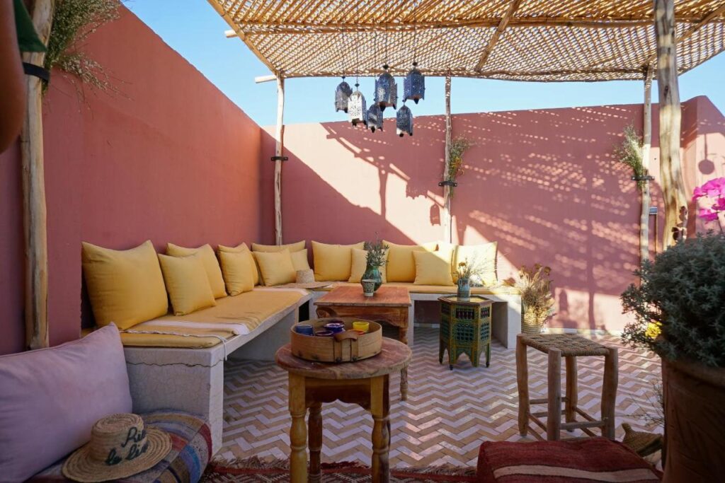 12 Charming Budget Riads in Marrakech 33 A cozy outdoor patio, reminiscent of budget riads in Marrakech, features a cushioned corner bench with yellow pillows, wooden tables, woven stools, potted plants, and a bamboo shade casting shadows against pink walls under a clear blue sky.