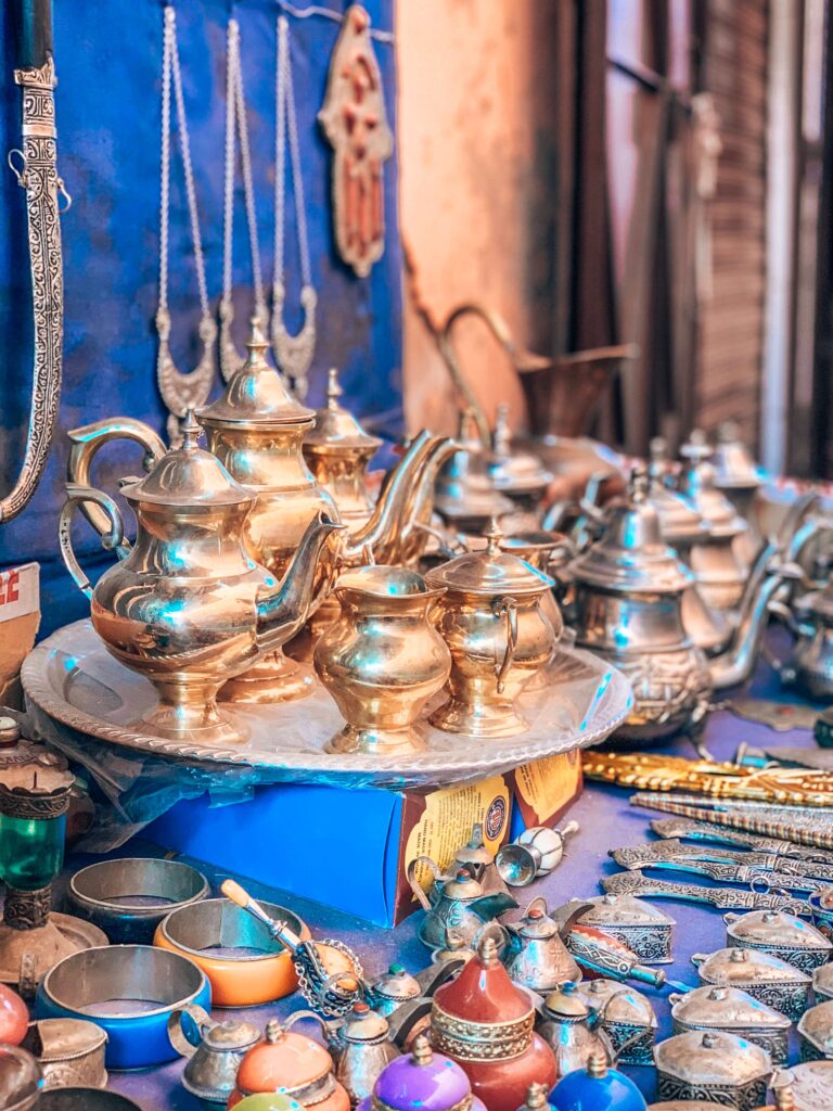 10 Unique Riads in Marrakech with Pool 44 A market stall displays shiny brass and silver teapots, cups, jewelry, and colorful trinkets on a blue tablecloth—typical treasures you might find while exploring Marrakech’s lively souks near budget riads in Marrakech.