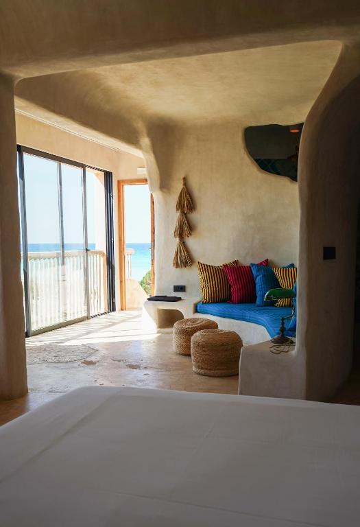 A cozy, sunlit room with curved beige walls features colorful pillows on a blue cushioned bench, two woven poufs, and sliding glass doors opening to a balcony with an ocean view—echoing the charm of boutique hotels in Formentera.