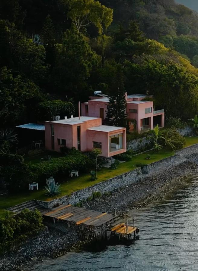 8 UNIQUE Places Where to Stay in Lake Atitlán 8 A modern pink house with large windows sits beside a lake, surrounded by lush trees and greenery—an ideal choice for those seeking where to stay in Lake Atitlan. A wooden dock extends into the water, with outdoor furniture on the grassy lawn near the shore.