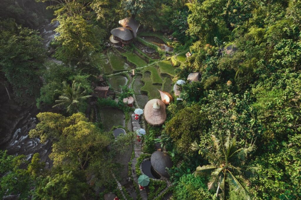 13 Most UNIQUE Places to Stay in Ubud 12 Aerial view of eco-friendly huts with thatched roofs nestled in lush green forest, terraced gardens, palm trees, and a river tracing the tranquil, tropical landscape—one of the most unique places to stay in Ubud.