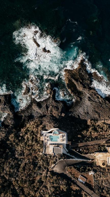 11 UNIQUE Boutique Hotels in Tenerife 24 Aerial view of a modern white house with a pool near a rocky coastline, waves crashing against the shore, and surrounding natural landscape—reminiscent of boutique hotels in Tenerife.