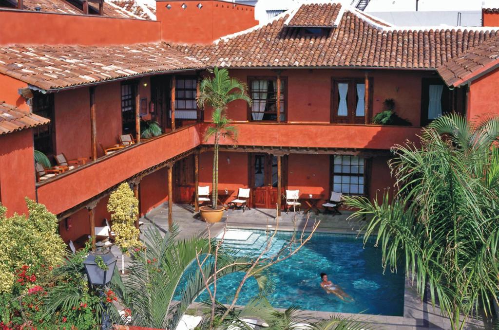 11 UNIQUE Boutique Hotels in Tenerife 34 A person swims in a pool surrounded by tropical plants in the courtyard of a two-story building with terracotta-colored walls and a tiled roof, capturing the relaxed charm of boutique hotels in Tenerife. Lounge chairs and balconies overlook the pool area.