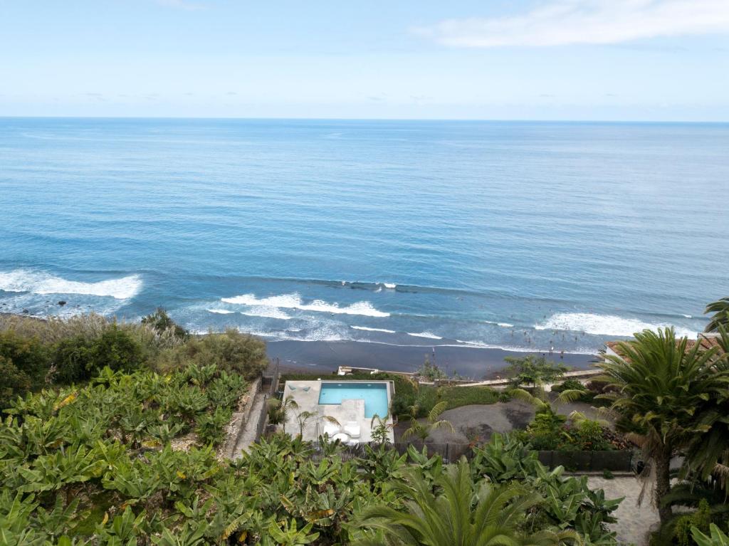 11 UNIQUE Boutique Hotels in Tenerife 39 A coastal view with lush green foliage in the foreground, a rectangular swimming pool below—like those at boutique hotels in Tenerife—and gentle waves rolling onto a dark sandy beach, meeting the expansive blue ocean under a partly cloudy sky.