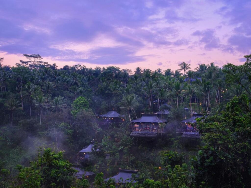 13 Most UNIQUE Places to Stay in Ubud 2 Misty jungle landscape at dusk with lush green trees and several tent-like structures nestled among the foliage, softly illuminated by lights, under a purple and blue sky—one of the unique places to stay in Ubud.