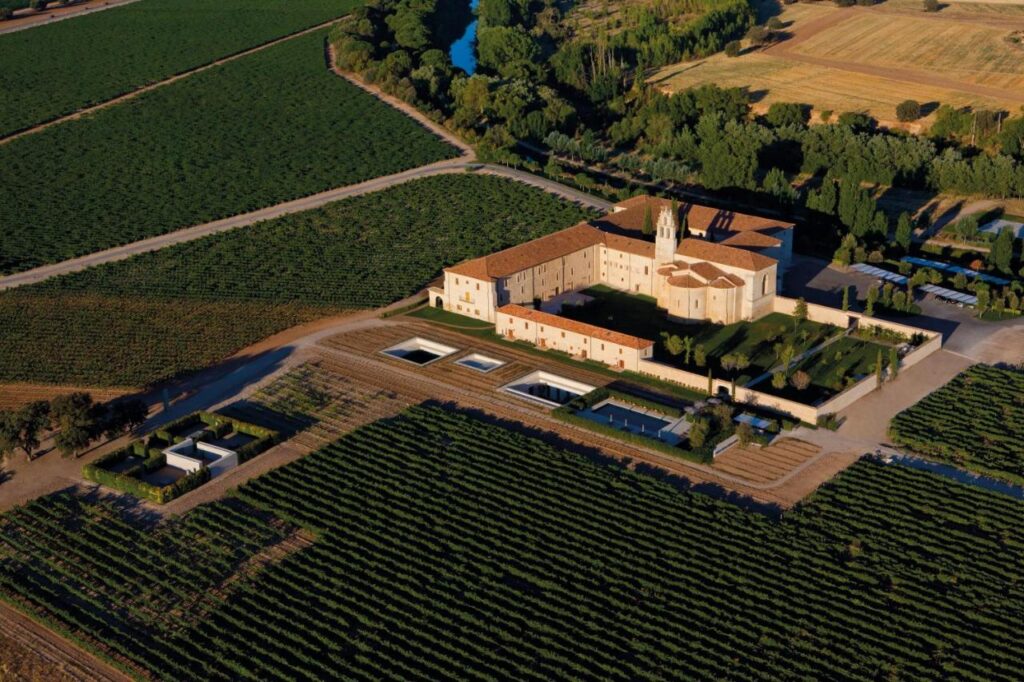 25 Weekend Trips from Madrid Max. 2h Away 42 Aerial view of a large historic building with a red-tiled roof, surrounded by vineyards and farmland—an ideal spot for weekend trips from Madrid. Geometric landscaping, small pools, and a scenic river complete the picturesque setting.