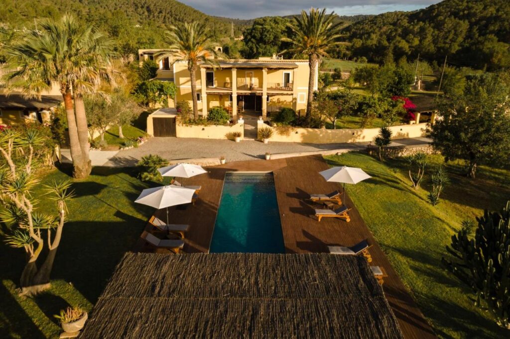 10 AUTHENTIC Finca Hotels in Ibiza 14 Aerial view of a luxury villa at one of the top finca hotels in Ibiza, featuring a rectangular pool, lounge chairs, lush lawn, palm trees, and scenic hills under a partly cloudy sky.