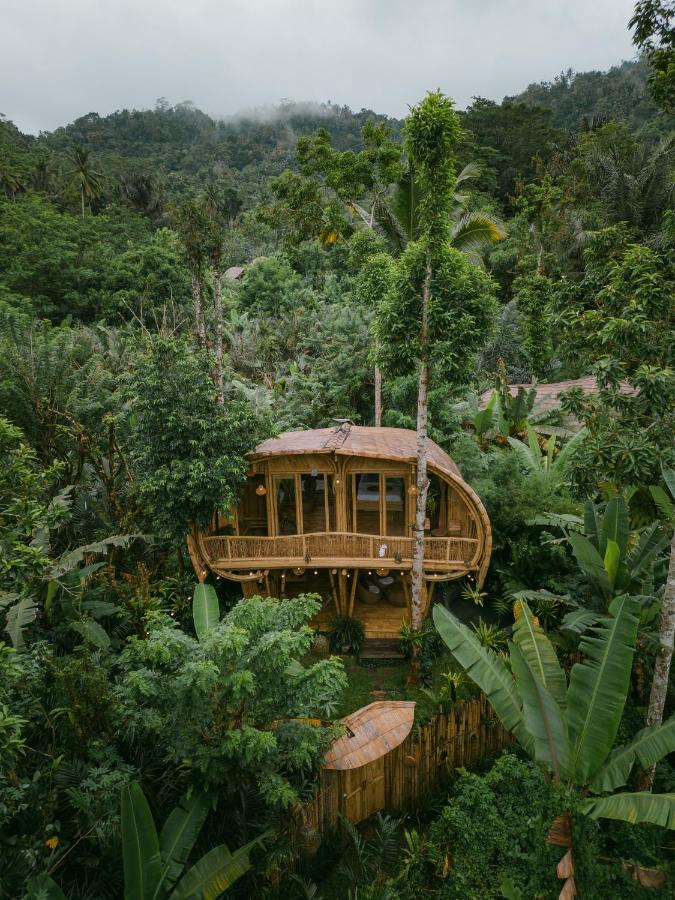 A bamboo treehouse with a curved balcony is nestled among dense tropical greenery and tall trees, offering one of the best family villas in Bali, surrounded by lush jungle and misty hills in the background.