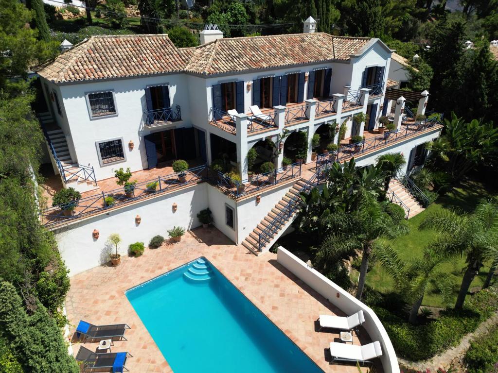 Adults-Only Hotels in Costa del Sol 20 A large Mediterranean-style villa with white walls, terracotta roof, multiple balconies, and staircases. Reminiscent of luxurious adult only hotels in Costa del Sol, it overlooks a rectangular pool surrounded by sun loungers and lush greenery.