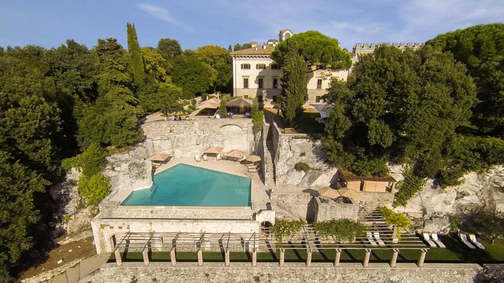 7 Best Agriturismos in Tuscany 23 Aerial view of a luxurious stone villa surrounded by lush trees, reminiscent of the finest agriturismos in Tuscany, featuring a terraced outdoor area with a rectangular infinity pool, lounge chairs, and shaded cabanas set against a hillside.