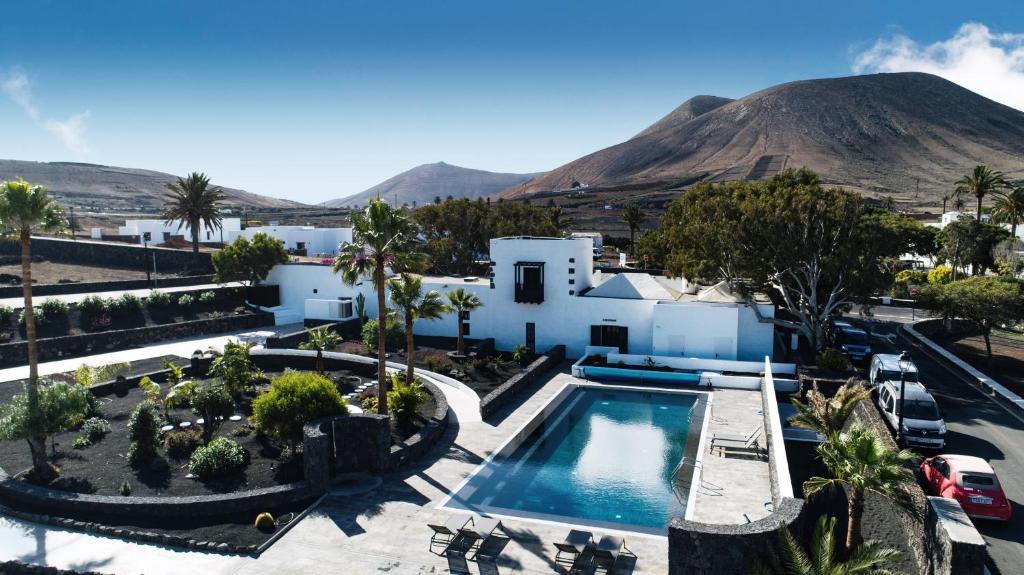 Unique Adults Only Hotels in Lanzarote 23 A modern white villa at an Adults Only Hotel in Lanzarote, featuring a large swimming pool, palm trees, and gardens, set against a volcanic landscape and clear blue sky. Several cars are parked along the driveway.