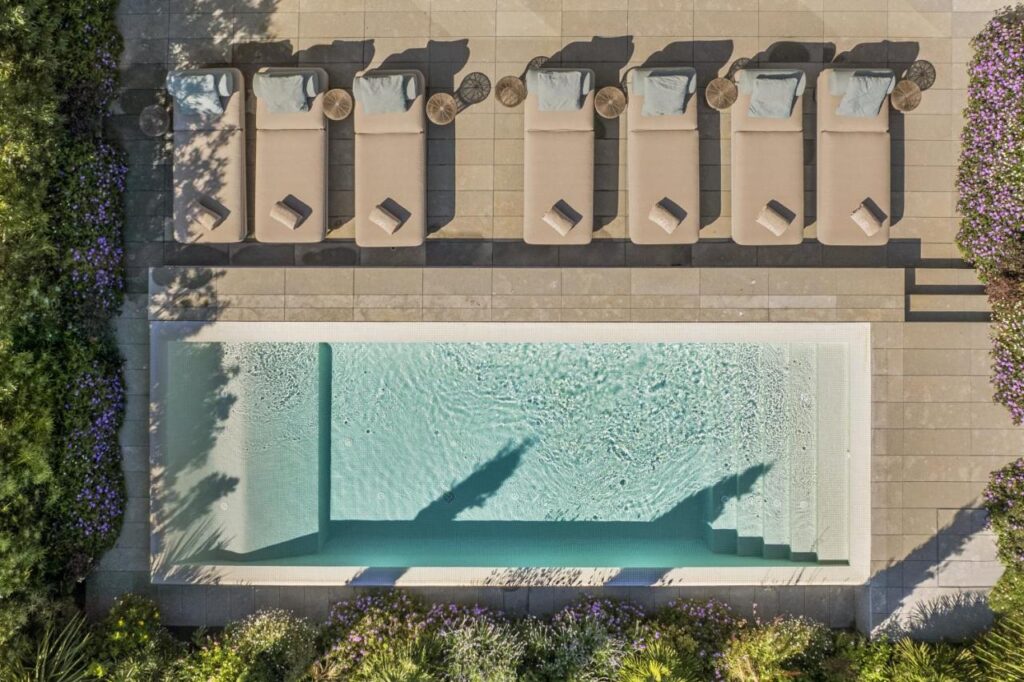 Top Barcelona Hotels with Rooftop Pool 9 Aerial view of a rectangular swimming pool with steps, surrounded by six beige lounge chairs and small round tables, bordered by greenery and purple flowers on a stone patio—an inviting scene found at many Barcelona hotels with rooftop pool amenities.