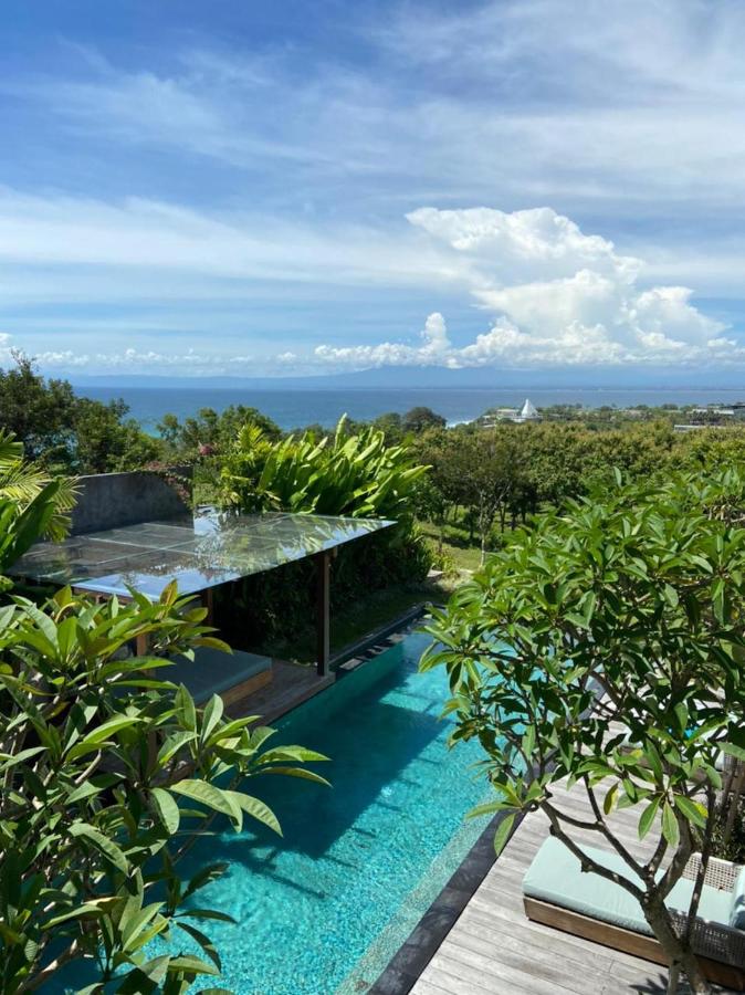 A sparkling blue pool surrounded by lush green trees and plants, with a wooden deck and lounge chair, overlooks a distant ocean—an idyllic scene at one of the best family villas in Bali under a partly cloudy sky.