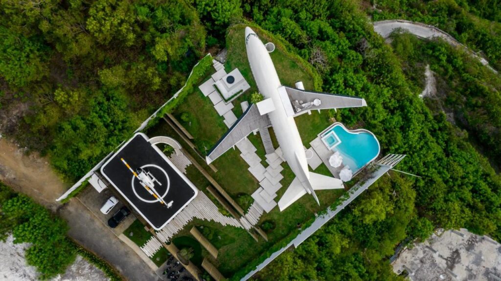 Aerial view of an airplane converted into a house, with a helipad and pool nearby, all surrounded by lush green trees and a winding road—an extraordinary retreat reminiscent of the best family villas in Bali.