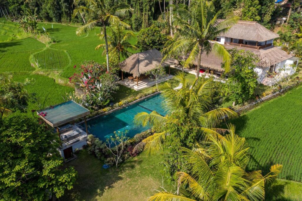 Aerial view of one of the best family villas in Bali, featuring thatched roofs, a large rectangular pool, and lush green rice fields surrounded by palm trees and dense vegetation.
