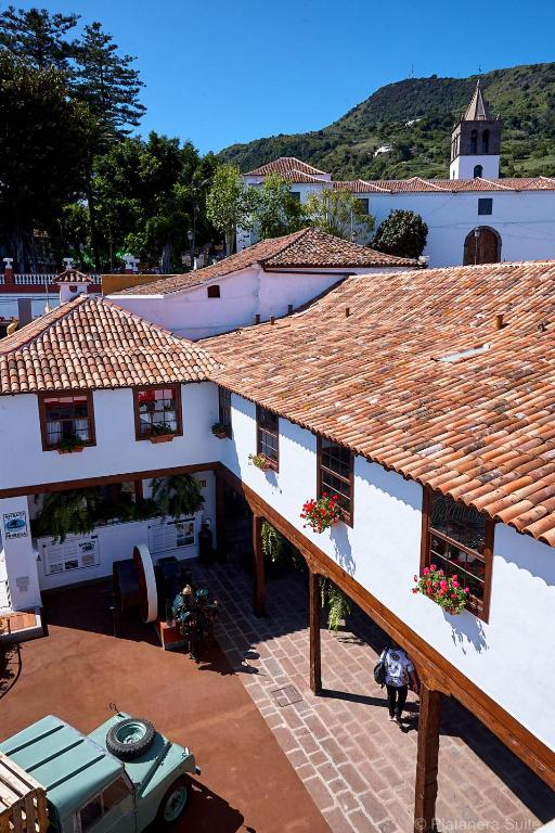 11 UNIQUE Boutique Hotels in Tenerife 28 A courtyard with white buildings and red-tiled roofs, flower boxes on windows, people strolling below, and a green vintage vehicle parked; mountains and a church tower rise in the background—capturing the charm of boutique hotels in Tenerife under a clear blue sky.