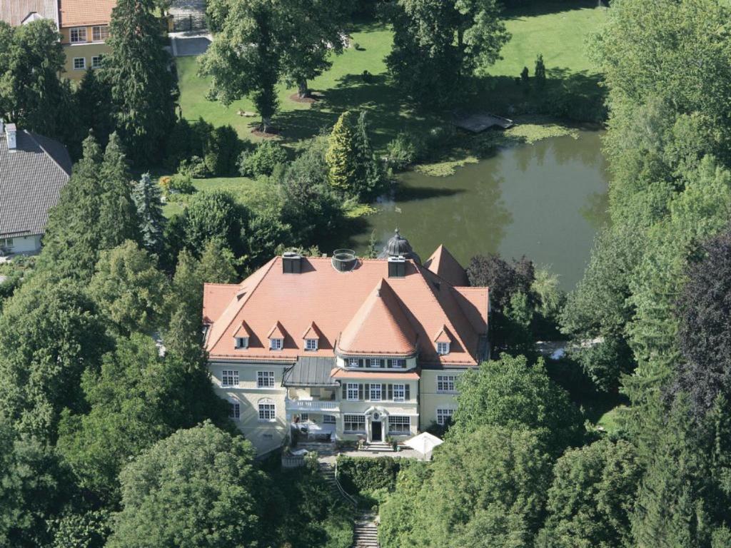 FAIRYTALE Castle Hotels in Bavaria 3 Aerial view of a grand mansion with a red-tiled roof, surrounded by lush green trees and gardens, reminiscent of the picturesque castle hotels in Bavaria, with a serene pond in the background.