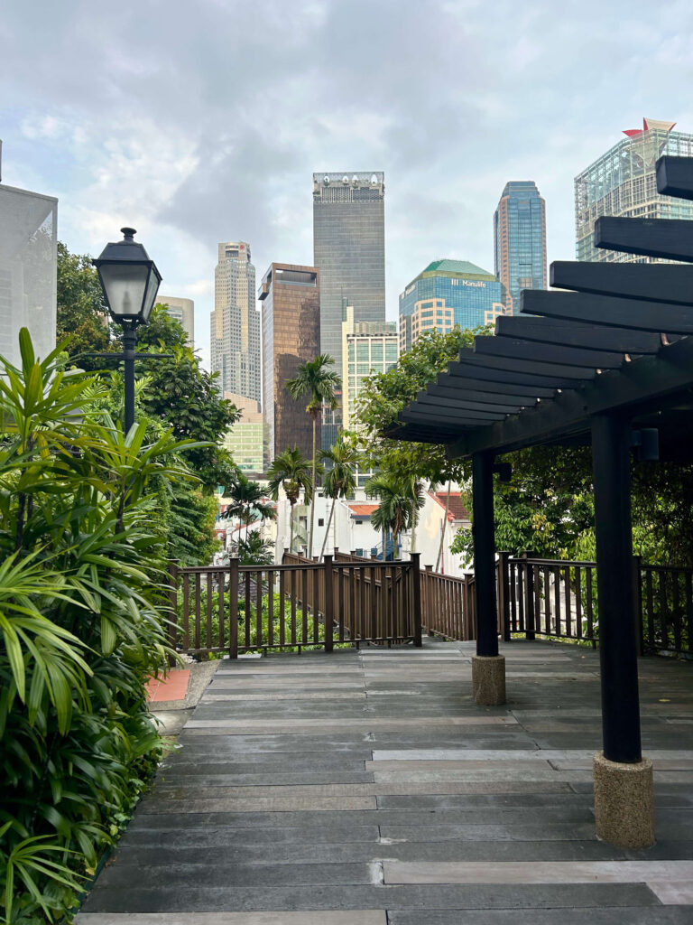 13 Most Unique Hotels in Singapore for an Incredible Holiday 59 A cityscape view featuring tall skyscrapers in the background, lush green plants, and unique hotels in Singapore, with a streetlamp and a shaded walkway with a wooden railing and pergola in the foreground under a cloudy sky.