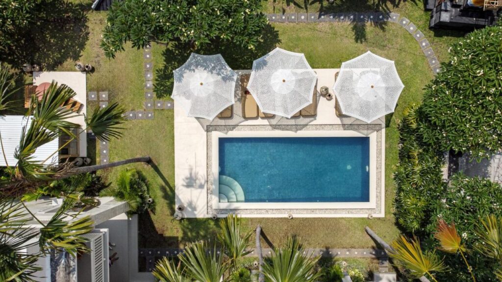 14 UNIQUE Boutique Hotels in Canggu, Bali 36 Aerial view of a rectangular swimming pool with three white umbrellas and lounge chairs beside it, surrounded by lush greenery—an inviting scene typical of boutique hotels in Canggu.