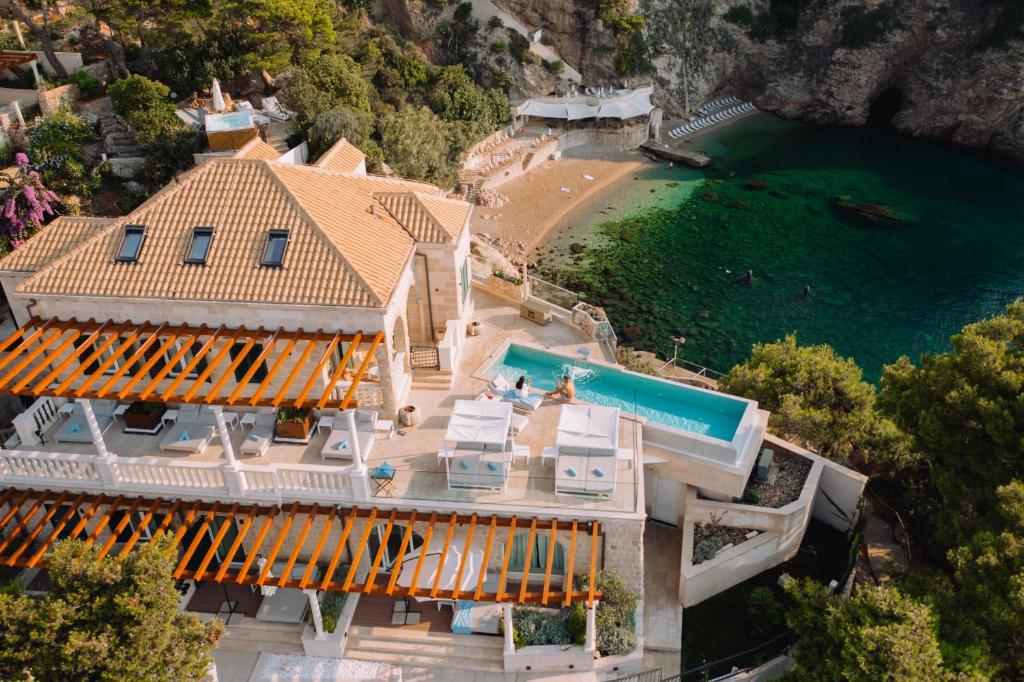 Most Unique Boutique Hotels in Dubrovnik 6 Aerial view of a luxurious seaside villa with a pool, sun loungers, and wooden pergola—reminiscent of the finest boutique hotels in Dubrovnik—overlooking a small sandy beach and turquoise cove surrounded by lush greenery and rocky cliffs.