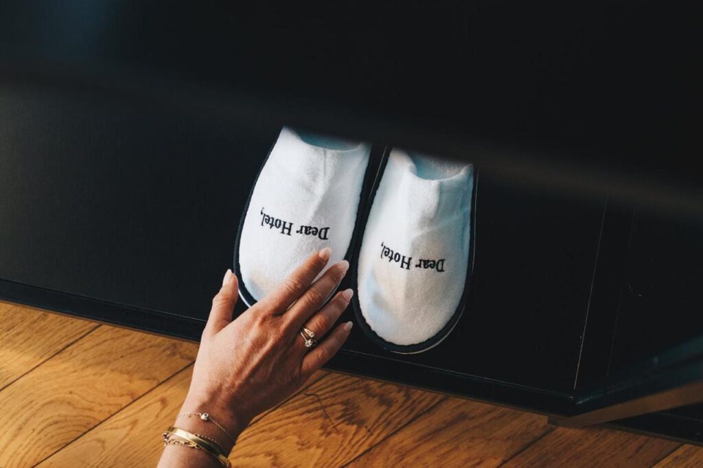 Where to Stay in Madrid: 14 Best Hotels 37 A hand reaches for a pair of white slippers with Dear hotel written on them, placed side by side on a dark surface above a wooden floor—perfect details for those wondering where to stay in Madrid first time.