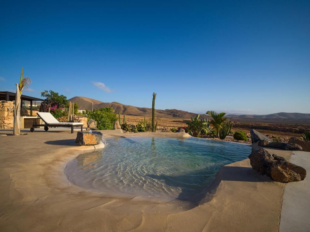 Adults Only Hotels in Fuerteventura 6 A small, natural-style pool with lounge chairs sits on a smooth stone patio, surrounded by desert plants and rocks—an oasis reminiscent of adults only hotels in Fuerteventura—overlooking arid hills under a clear blue sky.