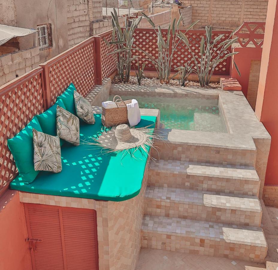12 Charming Budget Riads in Marrakech 32 A small rooftop patio in the style of budget riads in Marrakech features a sunken plunge pool, tiled steps, and a built-in bench with green cushions, tropical pillows, and touches like a straw hat, basket, and towels. Tall plants line the back fence.