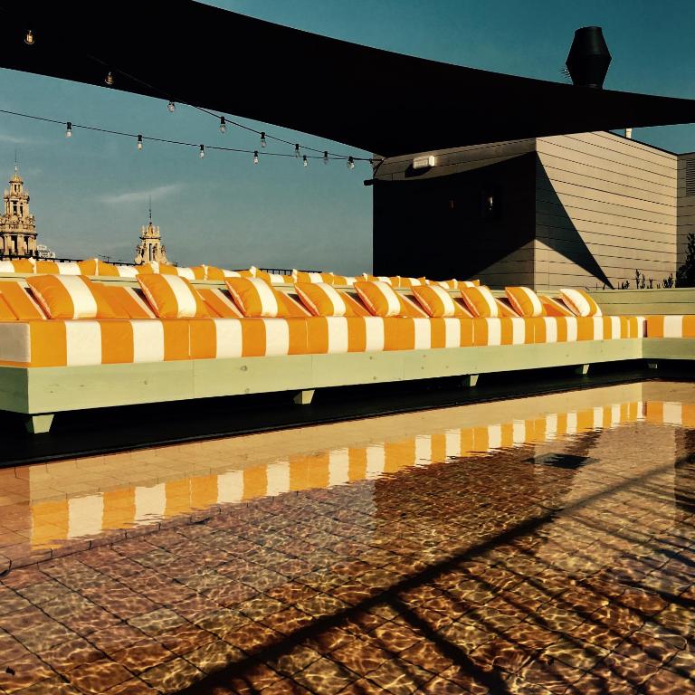 Top Barcelona Hotels with Rooftop Pool 42 A row of yellow and white striped lounge chairs sits by a shallow rooftop pool, with string lights overhead and city towers in the background—an inviting scene found at top Barcelona Hotels with Rooftop Pool under a clear sky.