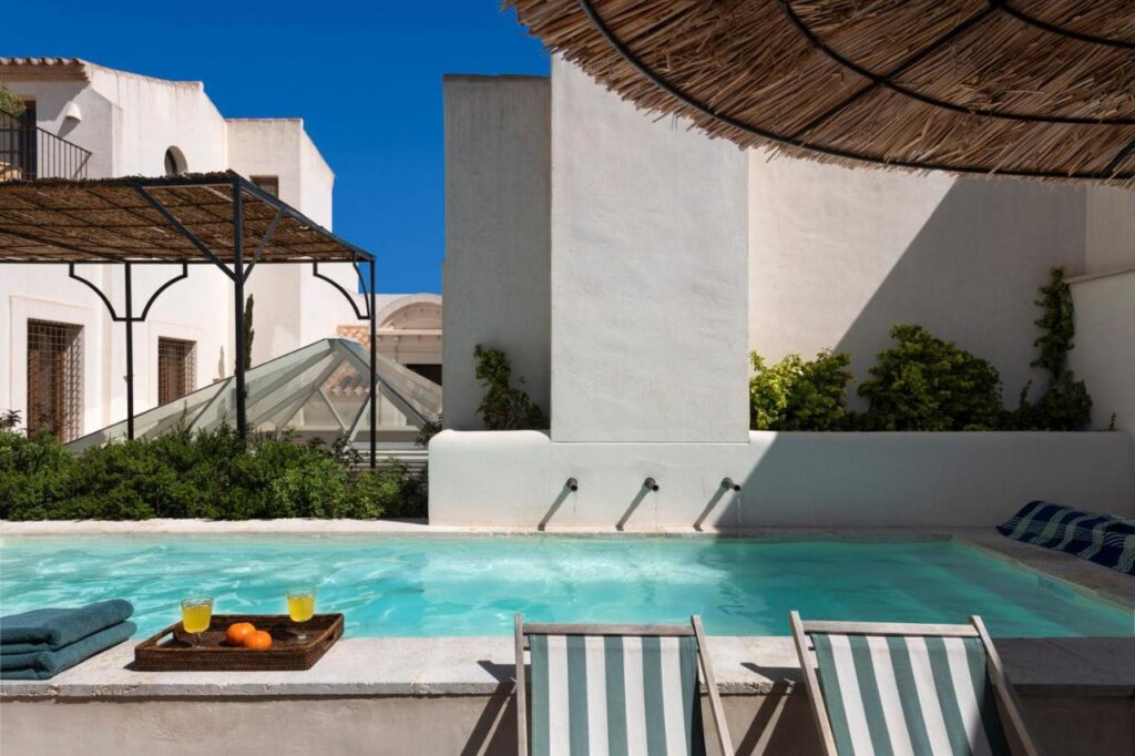 10 UNIQUE Hotels with Pool in Seville 8 A small outdoor pool with clear water is surrounded by white buildings, evoking the charm of hotels with pool in Seville. Two striped lounge chairs, fresh fruit, and green plants add to the relaxing, sunny atmosphere under a blue sky.