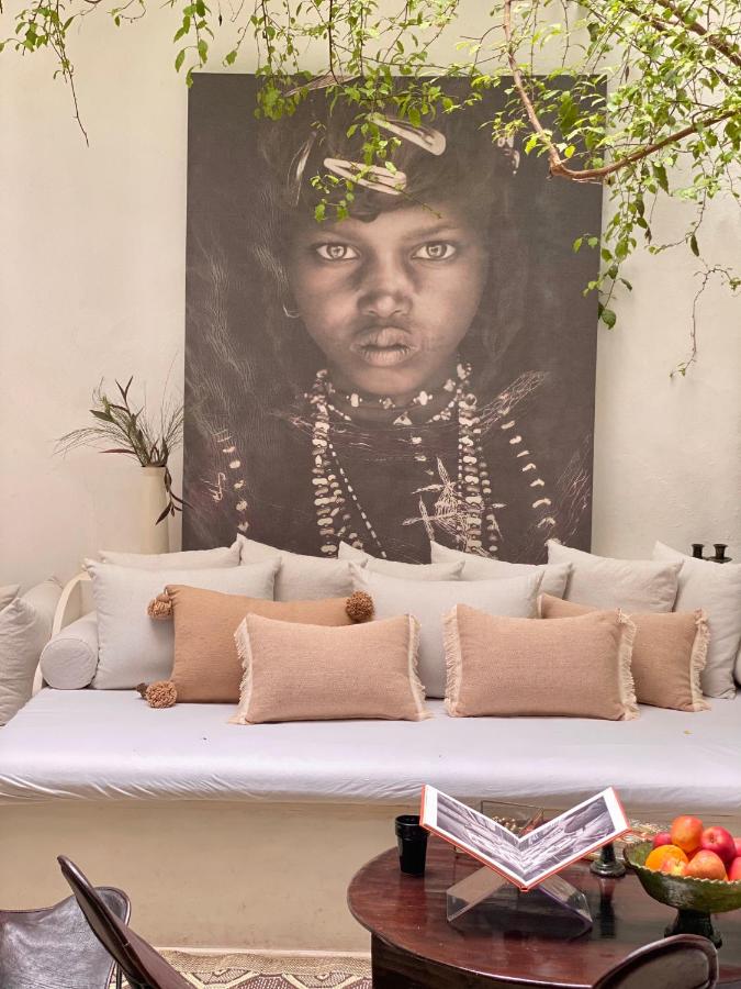 12 Charming Budget Riads in Marrakech 16 A cozy seating area, much like those found in budget riads in Marrakech, with a white cushioned bench, beige pillows, a round wooden table with fruit and an open book, and a striking black-and-white portrait of a child adorned with beads above the bench.