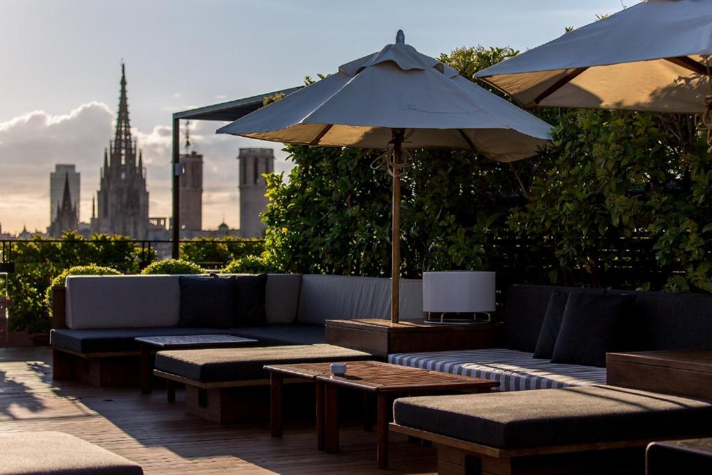 Top Barcelona Hotels with Rooftop Pool 37 A rooftop lounge with cushioned seating, wooden tables, and large umbrellas, surrounded by greenery—much like those found at Barcelona Hotels with Rooftop Pool. In the background, a city skyline and a prominent cathedral spire rise against a partly cloudy sky.