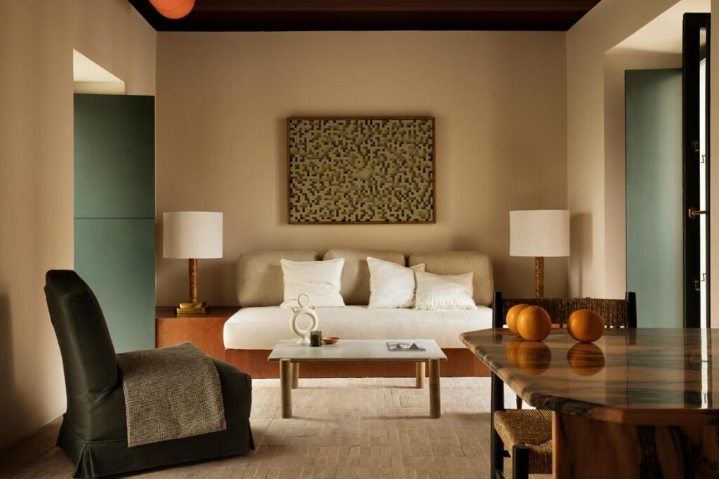 10 UNIQUE Hotels with Pool in Seville 40 A cozy living room with a cream sofa, neutral cushions, two white table lamps, modern abstract wall art—reminiscent of elegant hotels with pool in Seville—a dark armchair with throw, marble dining table with orange vases, and soft natural lighting.