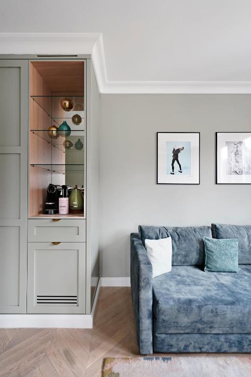 Unique Boutique Hotels in Salzburg 18 A cozy living room corner inspired by boutique hotels in Salzburg, with a blue sofa, colorful cushions, wall art, and a built-in cabinet displaying glassware, mugs, and coffee machines against light gray walls and wood flooring.