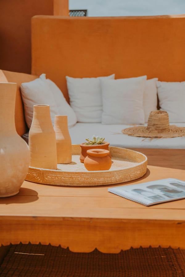 12 Charming Budget Riads in Marrakech 48 A cozy outdoor seating area, typical of budget riads in Marrakech, features white cushions, an orange wall, a wooden table with a woven tray holding clay and ceramic vases, a small potted succulent, and an open magazine.