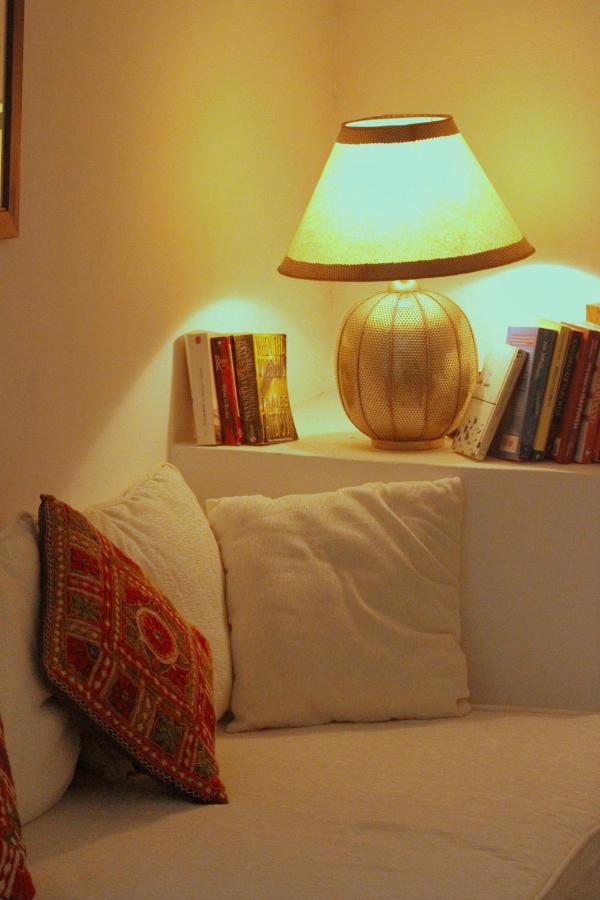 7 UNIQUE Riads in Fez You Won't Forget 25 A cozy corner inspired by riads in Fez, with a white sofa, colorful patterned cushion, and a lit table lamp on a shelf beside several books against a softly lit wall.