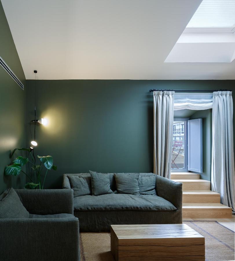 Best Boutique Hotels in San Sebastian 11 A modern living room with green walls, a grey sofa, and a wooden coffee table evokes the style of boutique hotels in San Sebastian. A potted plant, wall-mounted lamp, and light curtains complete the airy space filled with natural light.