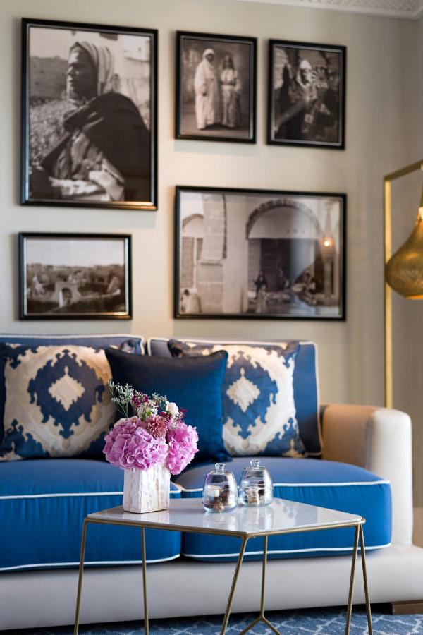 9 UNIQUE Riads in Tangier 8 A stylish living room, inspired by riads in Tangier, features a blue and white sofa with patterned pillows, a small table with two glasses and a vase of pink flowers, and black-and-white photographs in black frames on the wall behind.