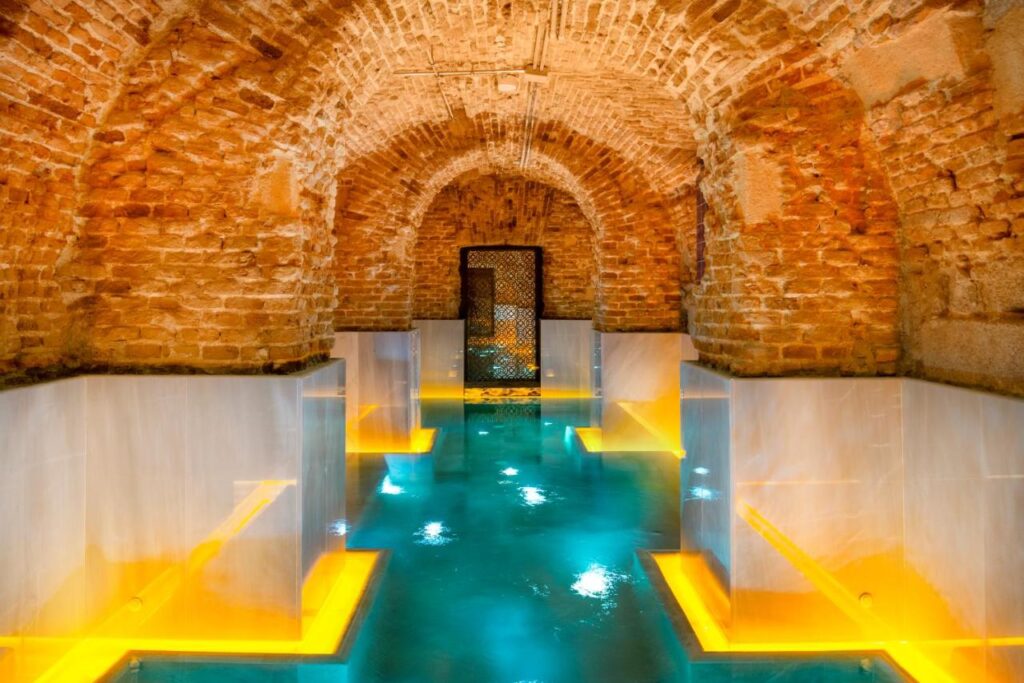 Where to Stay in Madrid: 14 Best Hotels 11 An underground spa with arched brick ceilings, warm yellow lighting, and turquoise water pools bordered by white marble—an ideal retreat for those wondering where to stay in Madrid first time, creating a serene and inviting atmosphere.