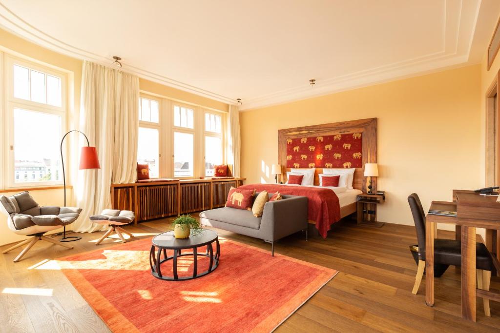 Boutique Hotels in Berlin Kreuzberg 14 A bright, spacious hotel room in one of the top boutique hotels in Berlin Kreuzberg, featuring large windows, a double bed with red bedding and elephant-patterned headboard, a gray sofa, armchairs, wooden desk, and coffee table on an orange rug.