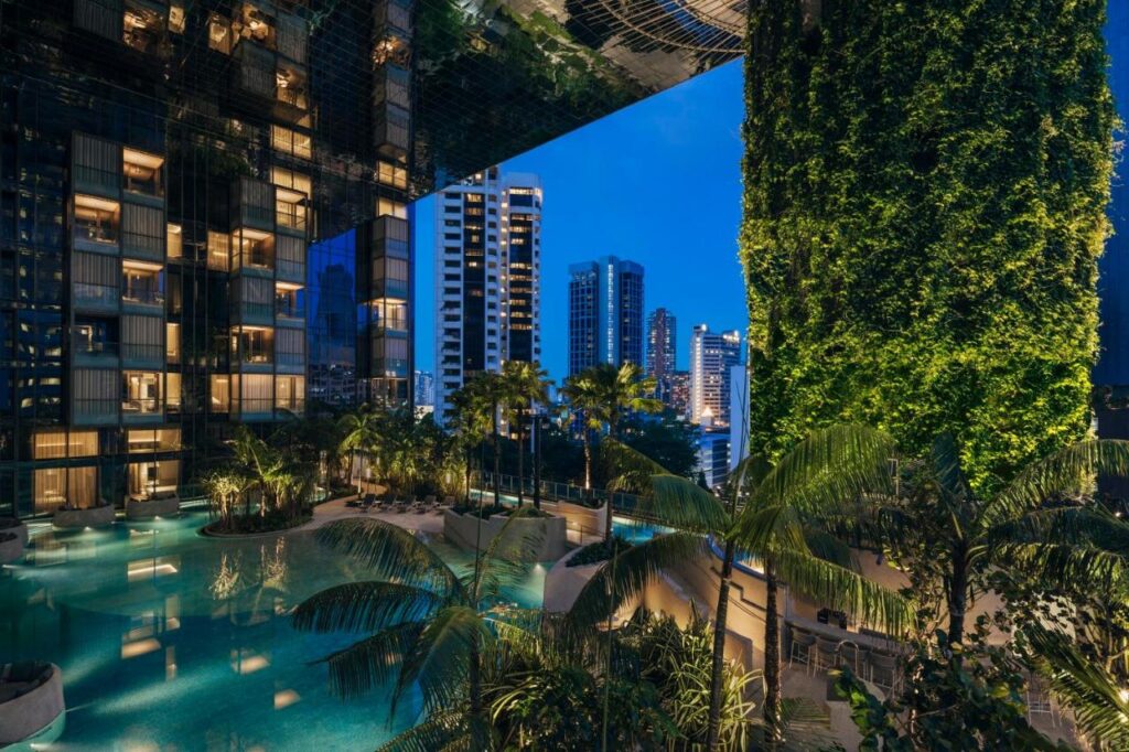 13 Most Unique Hotels in Singapore for an Incredible Holiday 40 Night view of a modern high-rise hotel with a lush green vertical garden, palm trees, and an illuminated swimming pool—one of the unique hotels in Singapore—with city skyscrapers visible in the background under a clear blue sky.