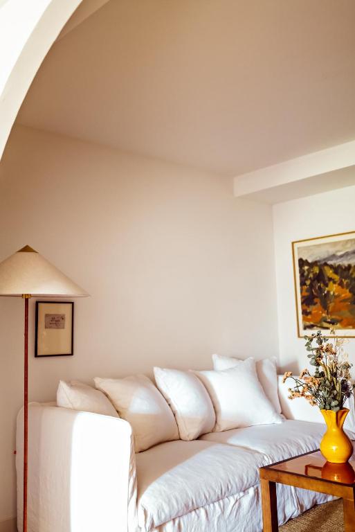 11 Unique Places to Stay in Provence 10 A cozy living room reminiscent of unique places to stay in Provence, with a white sofa, several white pillows, a wooden side table holding a yellow vase of flowers, a tall floor lamp, and framed artwork on plain white walls.