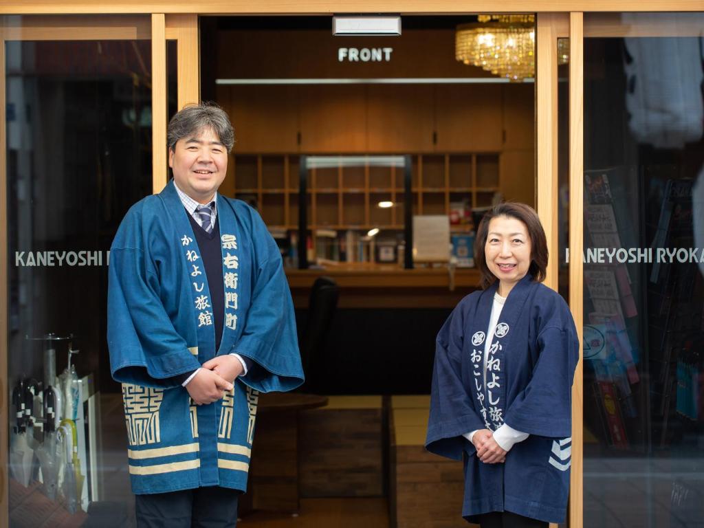 8 BEST Ryokans in Osaka 21 Two people wearing traditional Japanese happi coats stand smiling at the entrance of Kaneyoshi Ryokan, one of the welcoming ryokans in Osaka, with a wooden reception area and a “FRONT” sign visible behind them.