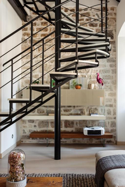 Where to Stay in Split: Hotel & Areas Guide 26 A modern black spiral staircase stands against a rustic stone wall, creating an inviting space that echoes the charm of Where to Stay in Split. Below, shelves display decorative items, a pink flamingo statue, plants, a printer, and glimpses of a sofa with a wooden coffee table.