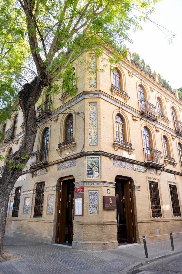 10 UNIQUE Hotels with Pool in Seville 13 A tan brick building on a street corner with decorative tiles and wrought iron balconies, reminiscent of some boutique hotels with pool in Seville; arched doorways and large windows adorn the shaded facade.