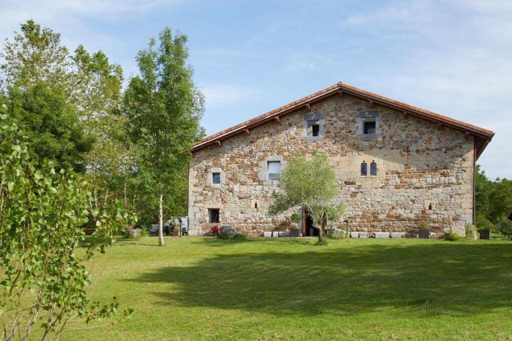 Best Boutique Hotels in San Sebastian 5 A rustic stone house with a sloped roof stands amid a grassy lawn and scattered trees, evoking the charm of boutique hotels in San Sebastian. The building features small windows and is surrounded by lush greenery under a partly cloudy sky.