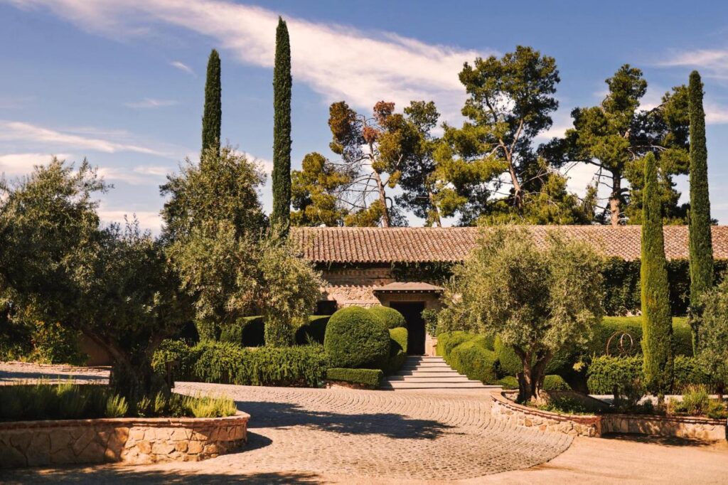 25 Weekend Trips from Madrid Max. 2h Away 86 A Mediterranean-style stone house with a tiled roof is surrounded by manicured hedges, tall cypress trees, olive trees, and a cobblestone driveway—an idyllic spot for weekend trips from Madrid under a clear blue sky.