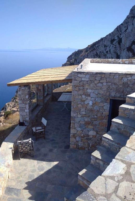 Authentic Hotels in Sikinos, Cyclades 6 A stone house with a patio overlooks the calm blue sea and rugged mountains—typical of charming hotels in Sikinos. Shaded areas under a wooden pergola and inviting patio furniture set the scene, while stone steps lead up from the tranquil terrace.